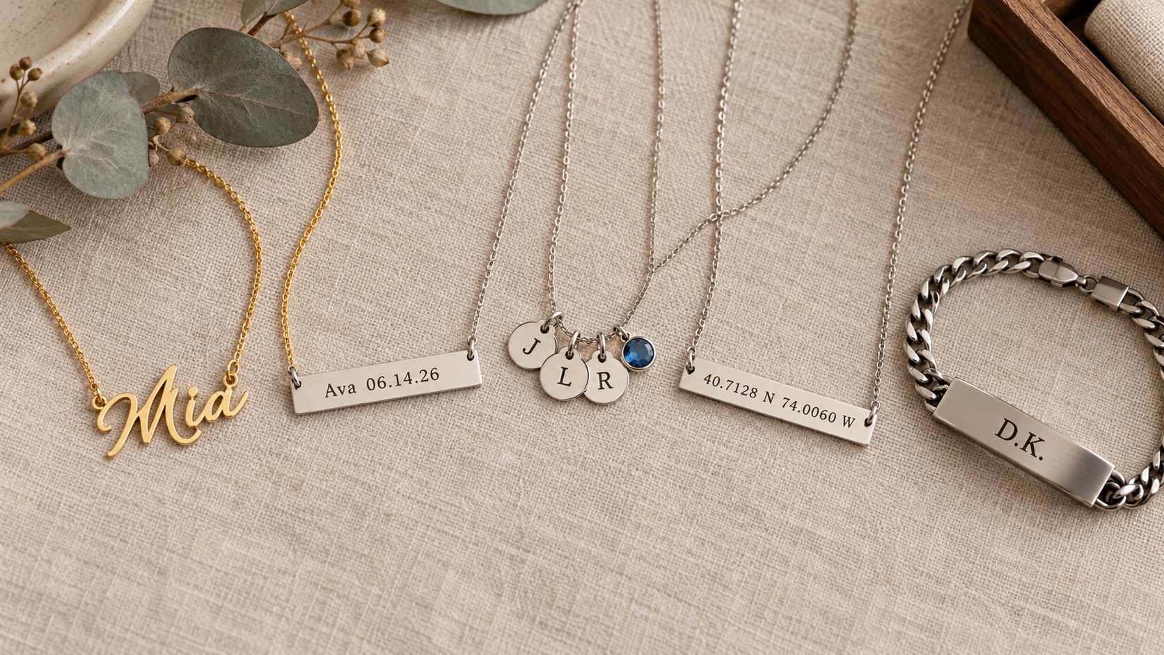 Flat lay of personalized jewellery types on linen: a gold name necklace reading Mia, an engraved silver bar, a birthstone disc necklace, a coordinates bar and a men's ID bracelet